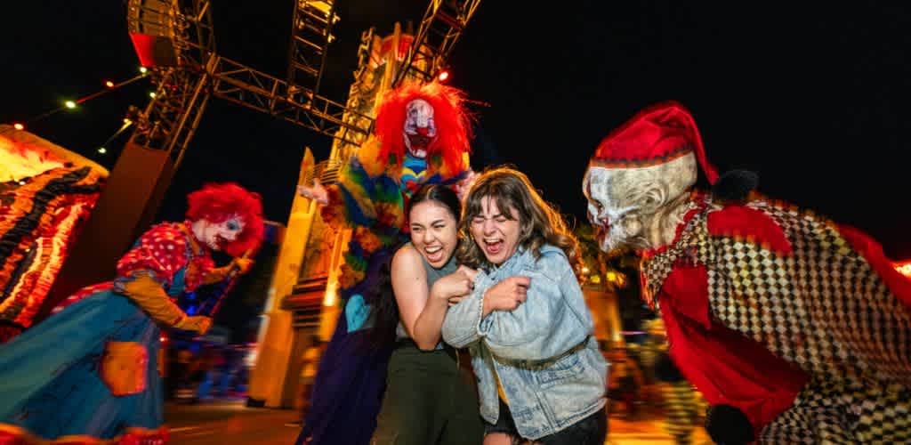 Two women laughing and hugging while surrounded by colorful clown and horror-themed figures at a carnival or amusement park nighttime event