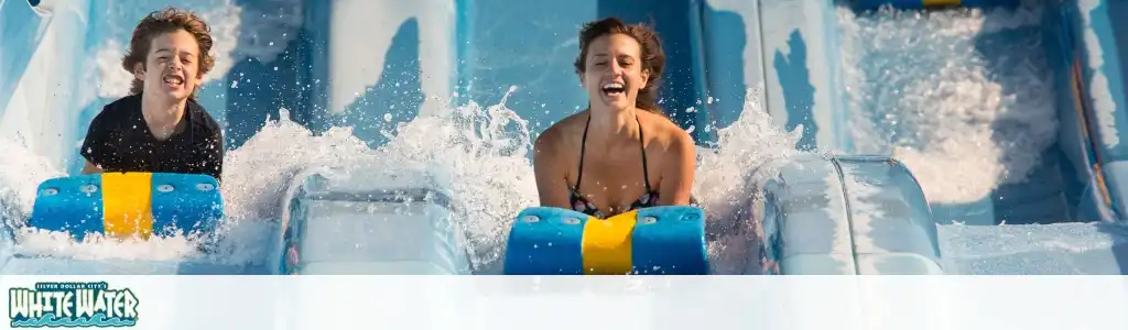 Two smiling people enjoy riding water slides at a water park with splashing water around them