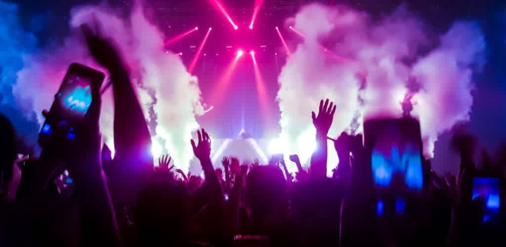 Crowd at a concert with raised hands and people taking photos, colorful lights and smoke effects creating an energetic atmosphere