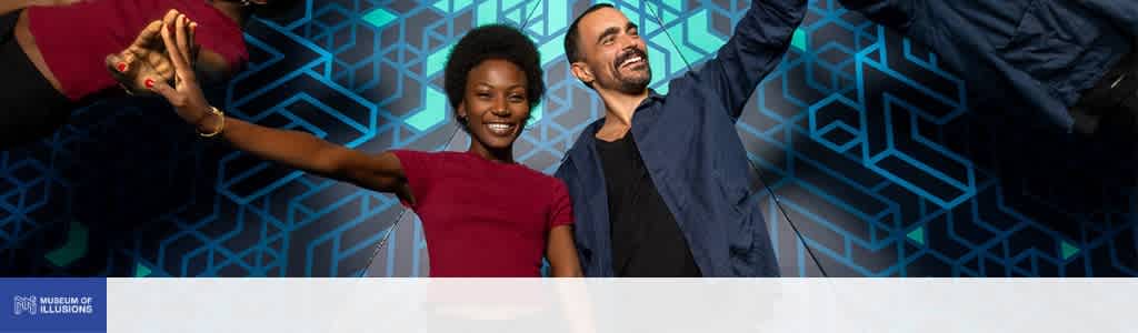 Two people smiling and dancing together in front of a blue geometric patterned background at the museum of illusions