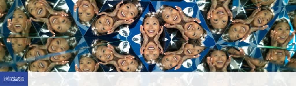 Multiple people smiling and looking excited inside a mirrored, kaleidoscope-like structure with reflections creating geometric patterns.