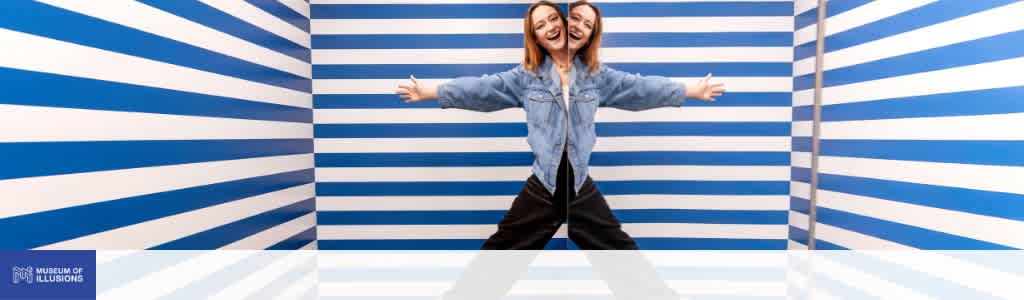 Two smiling women standing in an optical illusion room with blue and white horizontal stripes creating a mirror effect.