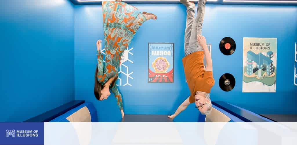 Two women are upside down in an illusion museum with blue walls and artwork, creating a playful and engaging interactive exhibit for visitors.