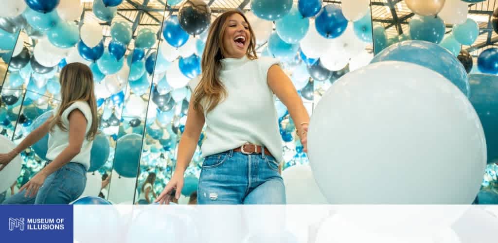 A woman smiling and laughing while surrounded by blue and white balloons in an immersive illusion exhibit at the Museum of Illusions