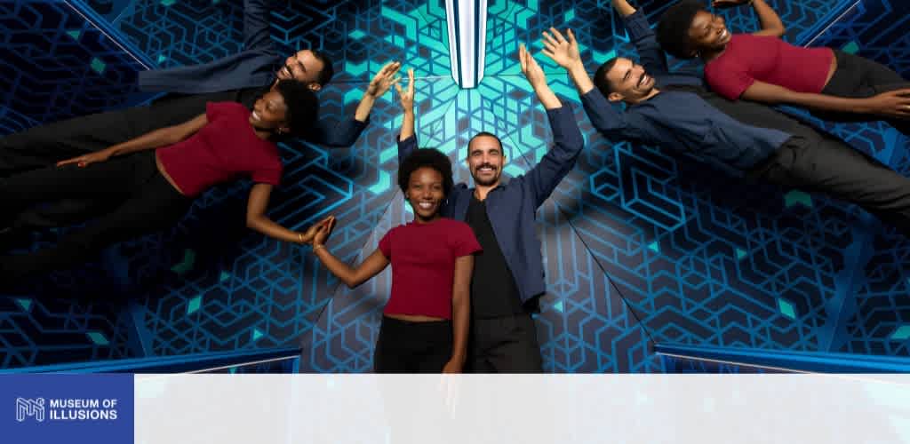 Four people are lying on a colorful geometric patterned floor with their heads together, smiling and raising their arms.