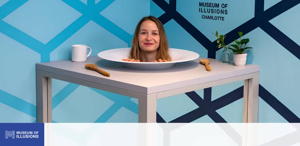 A woman with her head in a large bowl on a table at the Museum of Illusions Charlotte exhibits a playful illusion with a colorful geometric background.