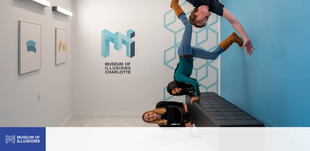 Two women and a man participate in a handstand near an art installation at Museum of Illusions Charlotte, with an abstract geometric mural and white walls.