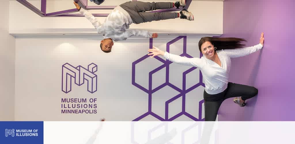 Two smiling women pose playfully against a purple and white Museum of Illusions Minneapolis wall with geometric design and logo.