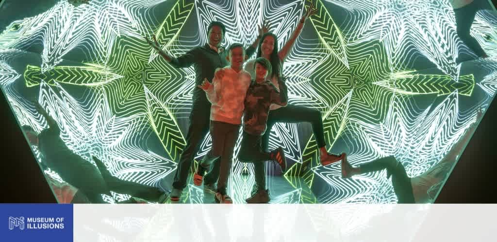 Four people pose in front of a vibrant, illuminated geometric pattern with green, black, and white lines at the Museum of Illusions.