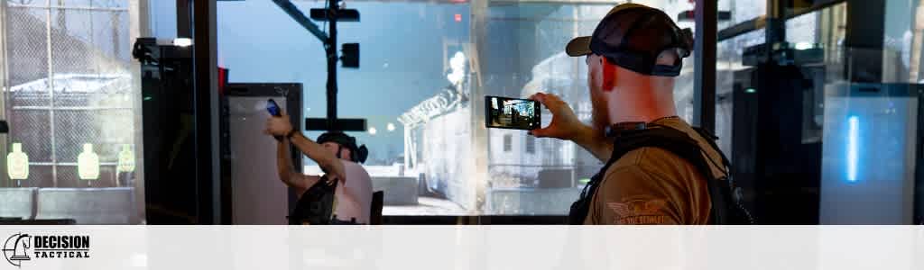 Two people are taking photos inside a modern indoor shooting range with glass targets and equipment.