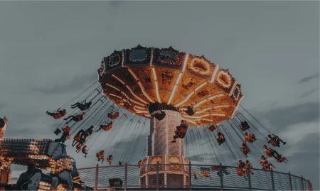 A brightly lit swinging amusement ride with multiple seats, rotating against a cloudy sky, at an outdoor fair or carnival setting.