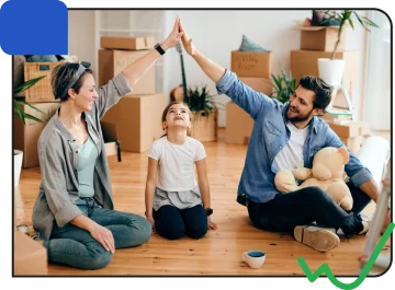 A family celebrating with high fives, sitting on the floor of a moving-in home surrounded by boxes and houseplants.