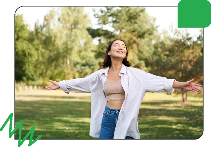 A woman standing outdoors with arms outstretched enjoying a sunny day in a park surrounded by trees