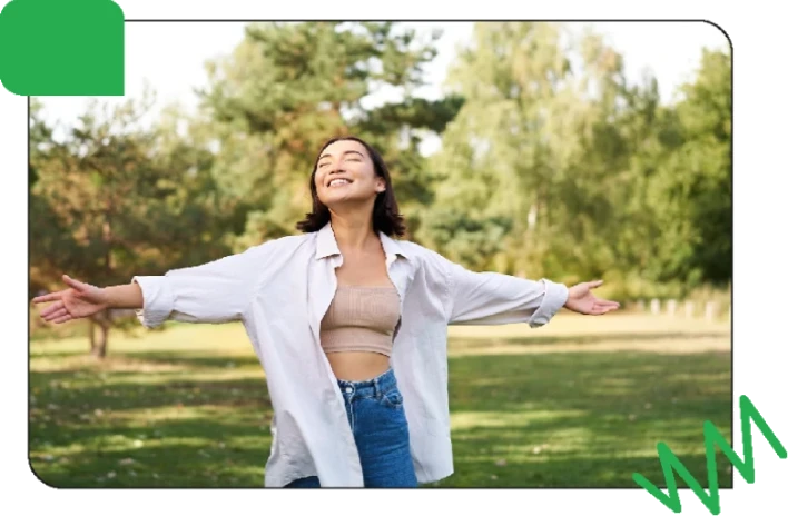A woman standing outdoors with arms outstretched enjoying a sunny day in a park surrounded by trees