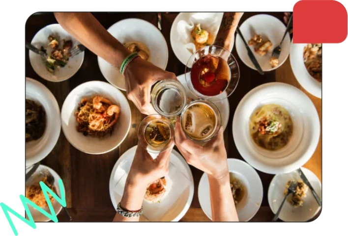 People clinking glasses of beer and cocktails over tables filled with various plates of food during a meal.